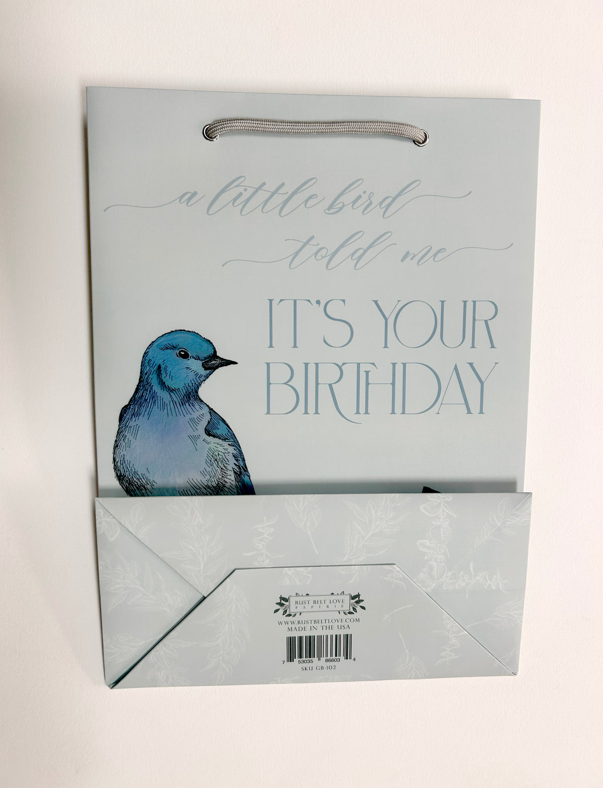 Flat photo of blue gift bag with florals that reads "A little bird told me it's your birthday" by Rust Belt Love. Bottom of bag is folded up to see the bottom packaging details