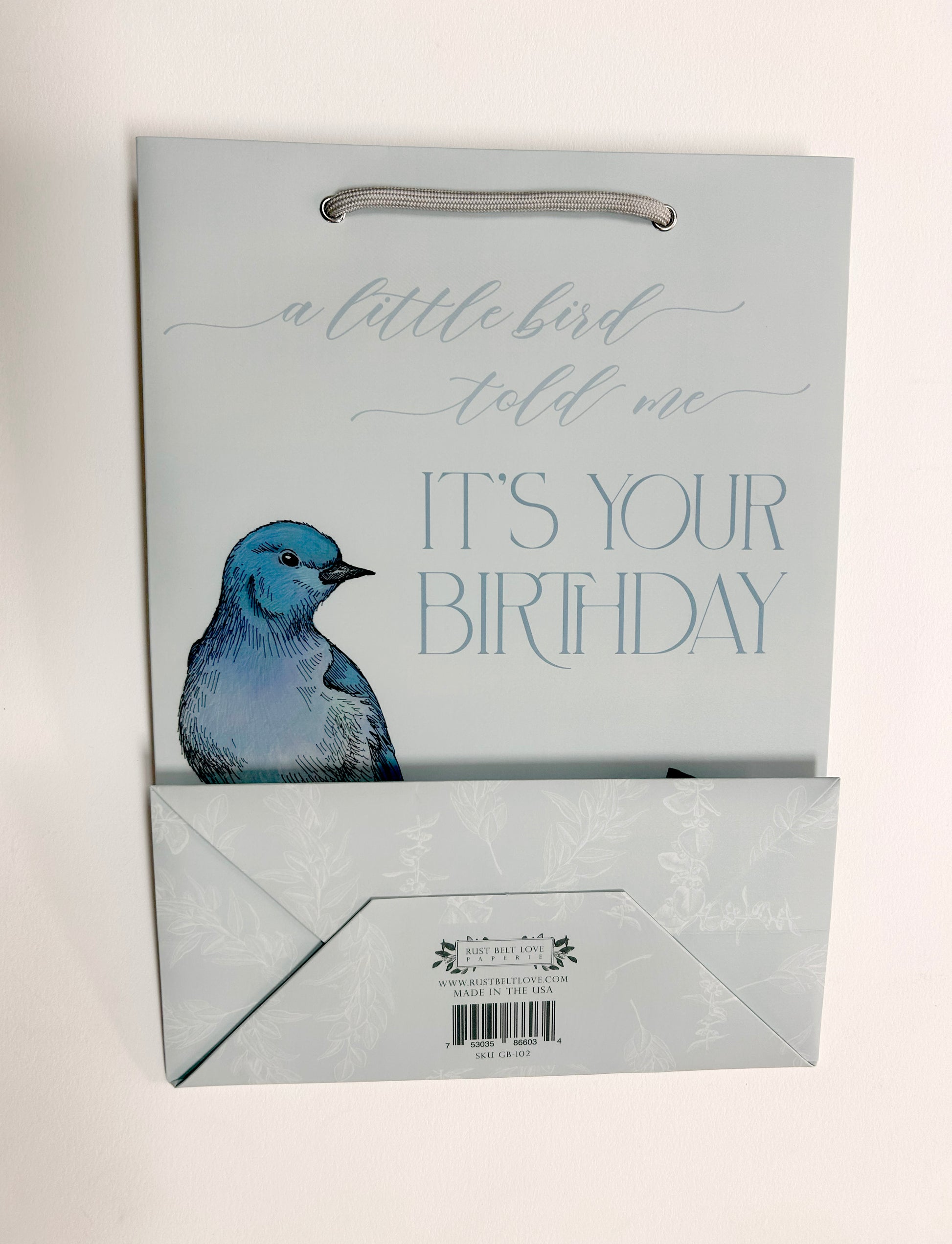 Flat photo of blue gift bag with florals that reads "A little bird told me it's your birthday" by Rust Belt Love. Bottom of bag is folded up to see the bottom packaging details